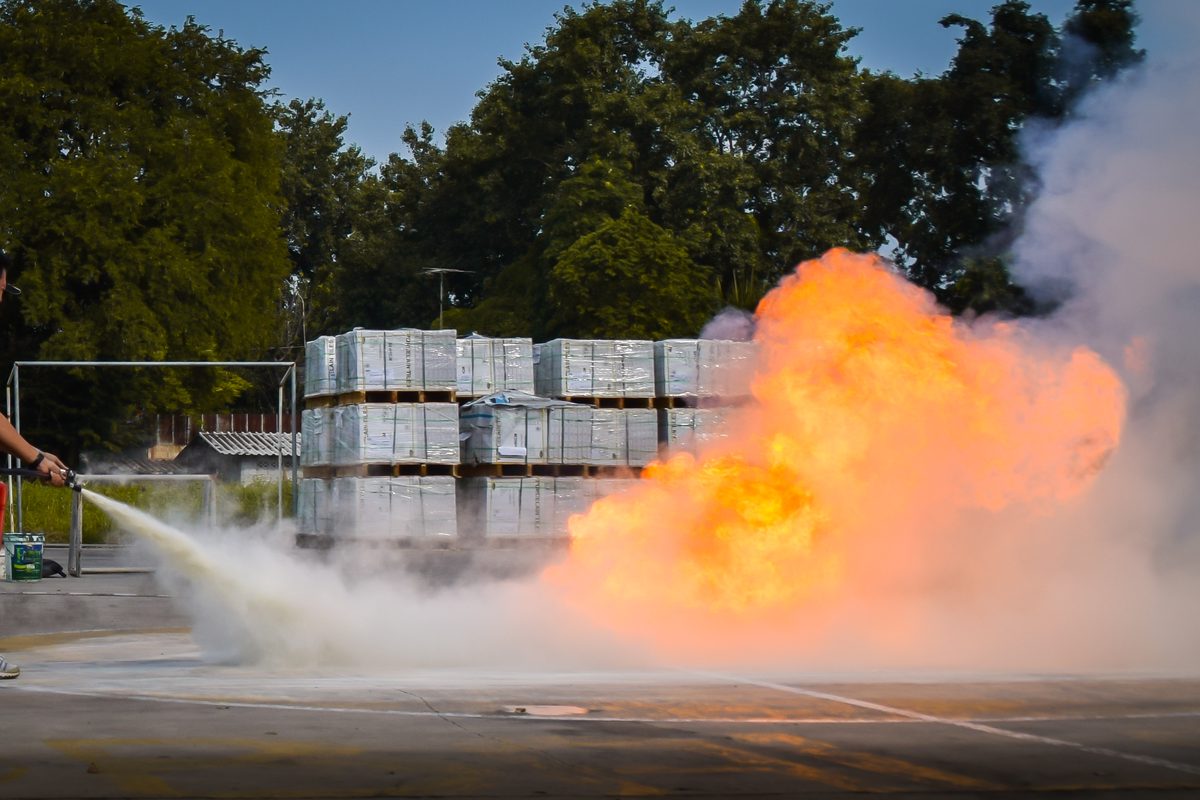 Industriearbeiter löscht Stichflamme an gestapelten Materialpaletten mit einem Schaumlöscher, Beispiel einer Gas- oder Staubexplosion in einem Lagerbereich